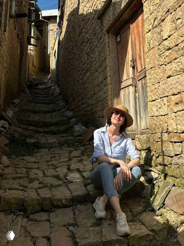 Узкая мощёная каменная улочка аула, высокие глинобитные стены и туристка в шляпе, сидящая у стены на солнце.