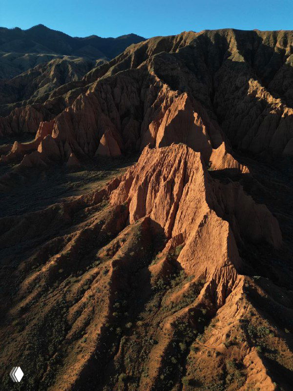 Красные каньоны Кыргызстана с глубокими складками и острыми хребтами на фоне синего неба, воздушная фотография подчёркивает марсианскую текстуру ландшафта.