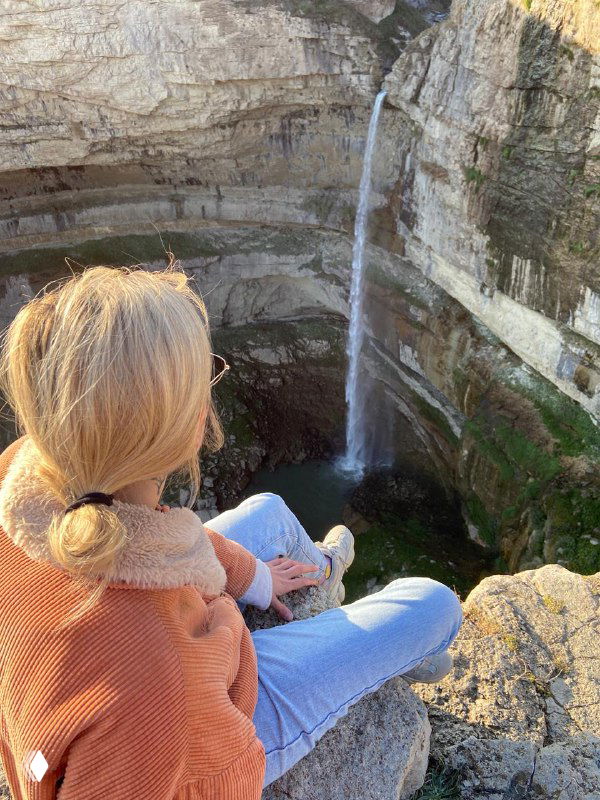 Водопад в горной котловине, рядом сидит девушка, смотрящая вниз на струи воды и обрывистую зелёную кромку ущелья.