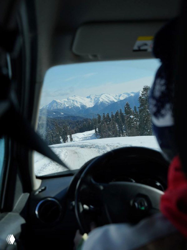 Вид из салона автомобиля на заснеженную дорогу и отдалённые горные пики, пассажир на переднем плане, путь к курортным зонам.