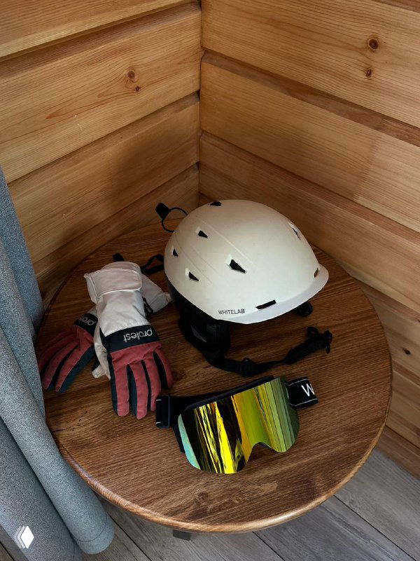 Круглый деревянный стол с горнолыжными аксессуарами: шлем, перчатки и зеркальные очки в уютном интерьере горной базы перед выездом.