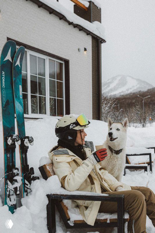 Отдыхающий в зимней одежде с хаски на шезлонге у домика, снег вокруг и уютная база отдыха на фоне склонов и деревьев.