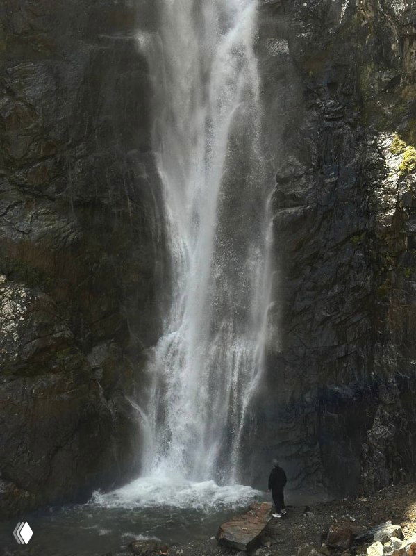 Высокий водопад в ущелье: вода стремительно стекает по скалам, у подножия виден человек для масштаба, окружение каменистое и зелёное.