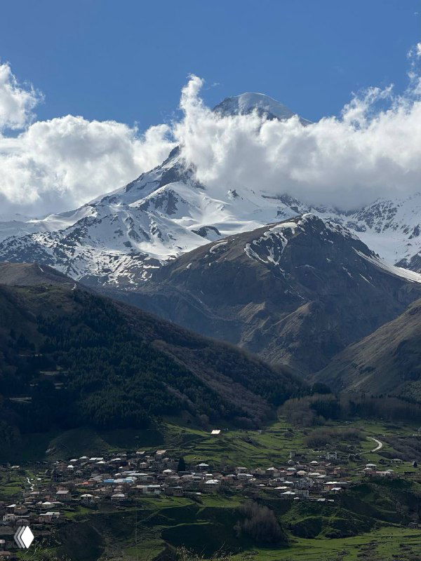 Заснеженная гора над деревней Степанцминда: панорама с домами в долине и крутыми склонами под голубым небом.
