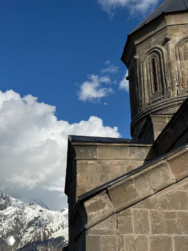 Каменная стена и купол грузинской церкви на фоне заснеженных гор и чистого неба — архитектурный фрагмент в горном пейзаже.