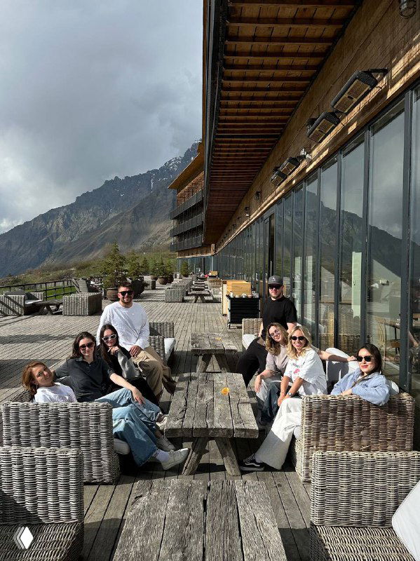 Гости на просторной террасе отеля: группа людей отдыхает на плетёной мебели с видом на горы и долину Степанцминды.