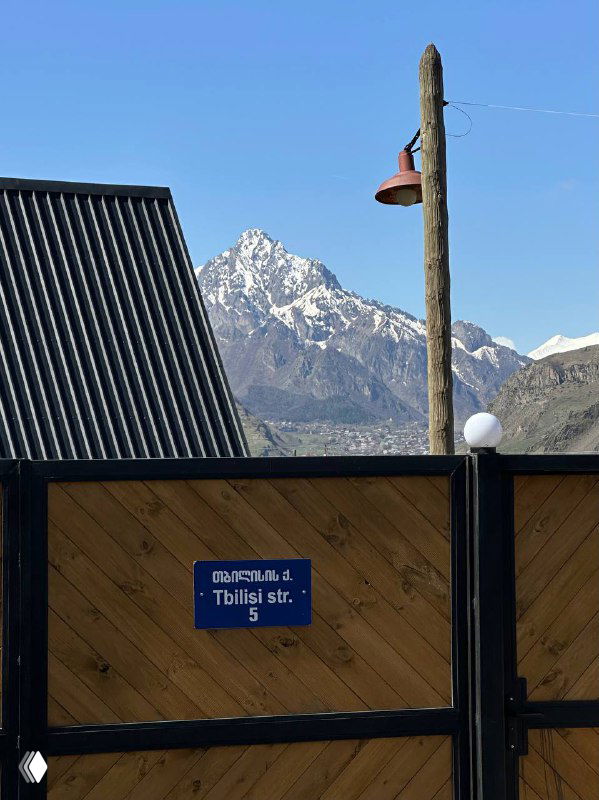 Деревянный забор с адресной табличкой «Tbilisi str.» на переднем плане и высокие вершины Кавказа на заднем плане под облачным небом.