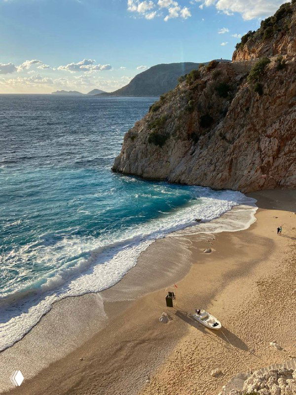 Пустынный галечный пляж у крутого скального берега, бирюзовая вода и небольшая лодка на песке — типичный вид средиземноморского побережья.