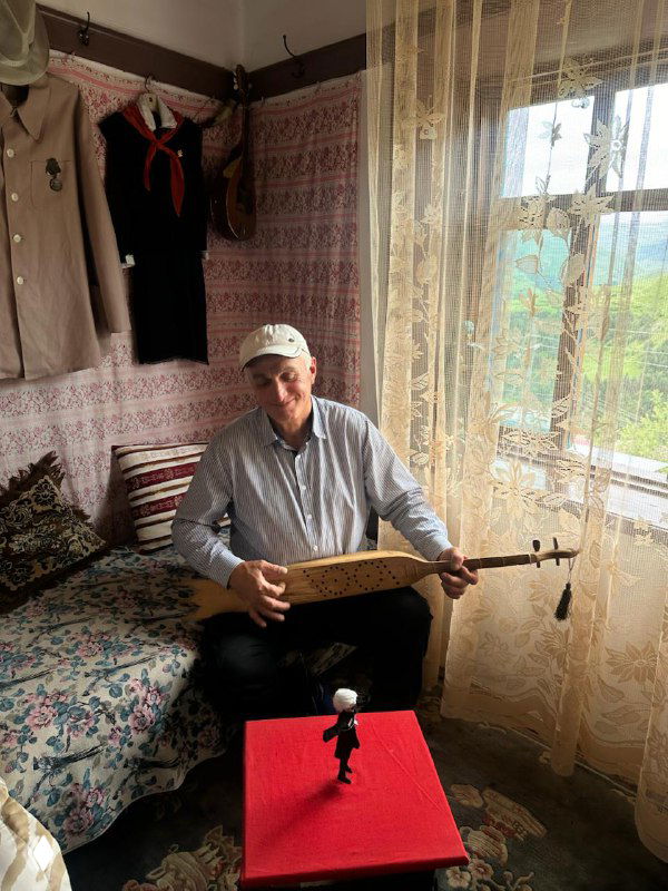 Местный музыкант в доме в ауле: мужчина с традиционным инструментом на фоне текстиля и уютной комнаты, живой быт гостей.