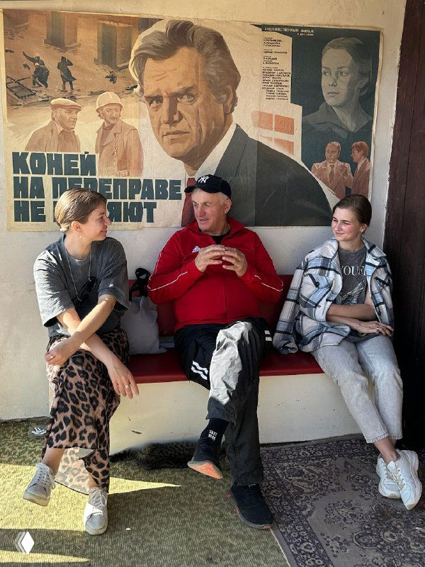 Группа людей на скамейке в ауле: разговоры местных и гостей, непринуждённая атмосфера и межкультурное общение в поездке.