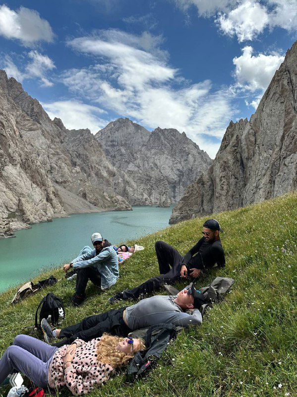 Группа туристов отдыхает на травяном склоне у бирюзового озера в ущелье Тянь‑Шаня; рюкзаки и спокойная горная панорама вокруг.