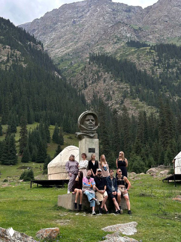 Группа путешественников у памятника на зелёной поляне рядом с юртами; на фоне — лесистые склоны и горные вершины, характерные для региона.