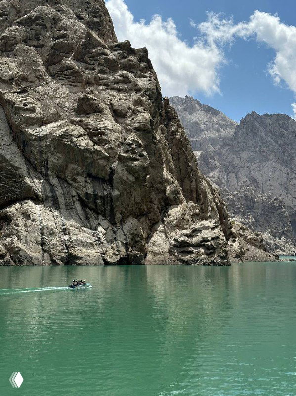 Бирюзовое горное озеро, окружённое отвесными скалами; спокойная гладь воды и маленькая лодка у подножия внушительных утёсов вдалеке.
