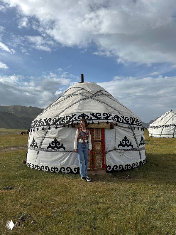 Традиционная белая юрта с декоративным орнаментом на степной поляне; турист у входа на фоне широкого горизонта и холмов Кыргызстана.