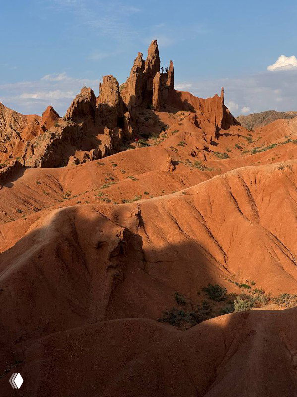 Высокие оранжево‑красные скалистые образования и колонны в полупустынном горном ландшафте Кыргызстана; выразительная геология и текстура пород.