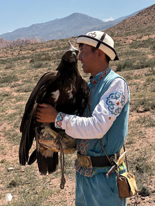 Местный сокольник в национальной одежде держит большого орла на руке на фоне засушливого горного ландшафта — традиционное ремесло и птица.