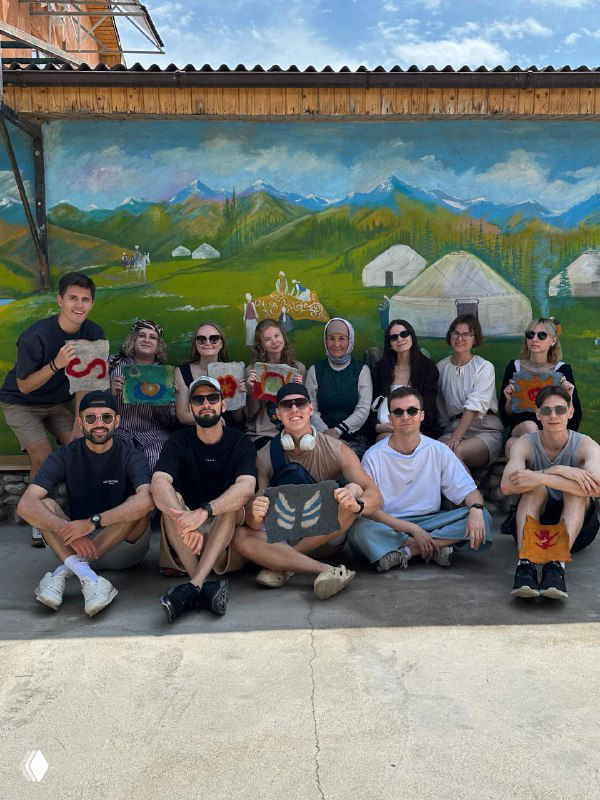 Группа путешественников на фоне раскрашенной стены с изображением гор и юрт — совместное фото на фоне местного арт-оформления в селе.