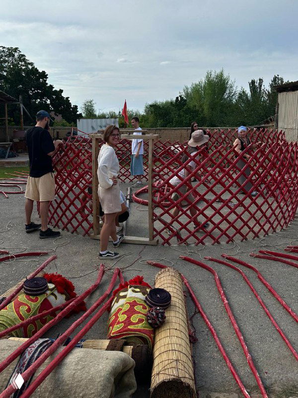 Сборка каркаса юрты: люди вокруг красной решётки-остова юрты, заготовки и полосы для её возведения на открытом пространстве.