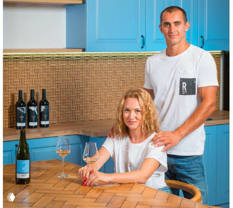 Фото двух людей за кухонным столом с бокалами и бутылками вина на фоне голубой кухни; атмосфера домашней дегустации и спокойного вечера