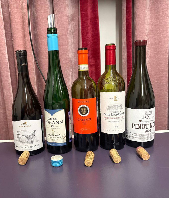 Несколько открытых бутылок вина разных форматов расположены на столе с корками и крышками — подборка для дегустации по эпохам.