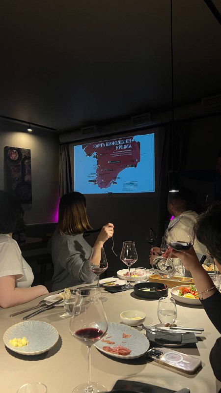 Участники дегустации за столом, видны бокалы и проекция на стене — атмосферный кадр с питерской встречи любителей крымских вин.