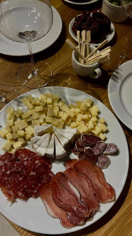 Тарелка с сырами и мясными нарезками на столе дегустации; бокал вина и столовые приборы создают атмосферу винного вечера.