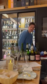 Сцена из зала дегустации: человек в голубом свитере рядом с бутылками и бокалами, на столе тарелки и аксессуары для оценки вин, динамика процесса.