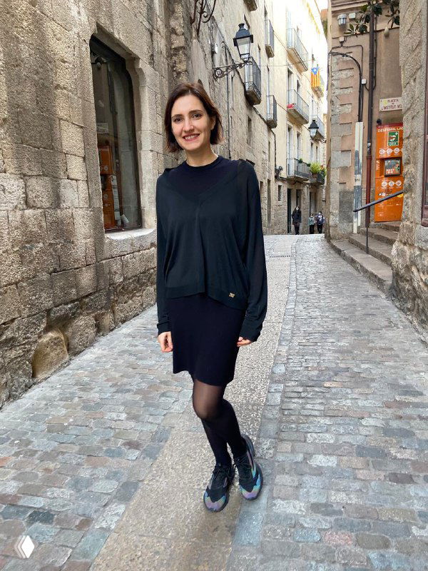 Женщина (Виктория Шешенина) стоит на мощёной узкой европейской улице в повседневной одежде, улыбается, видны фасады домов и фонарь.