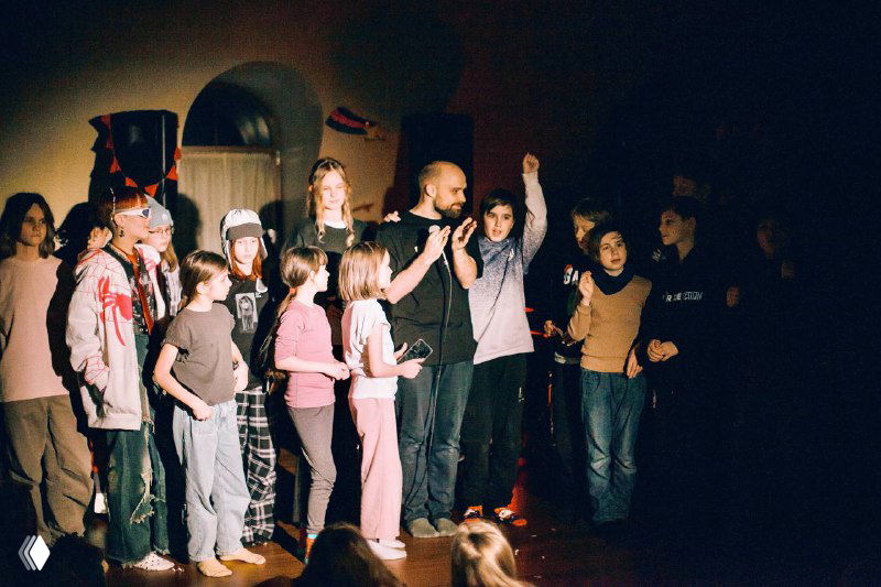 Группа детей и подростков на сцене под направленным светом: разные эмоции, взросление через коллективные активности и игры в помещении.