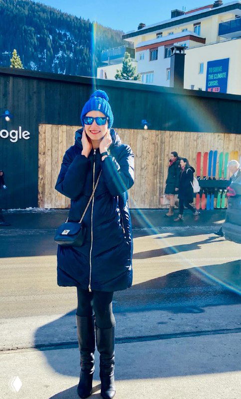 Женщина в тёмной зимней куртке и синей шапке стоит на солнечной улице Давоса; на заднем плане заснеженные горы, здания и уличные декорации международного форума.