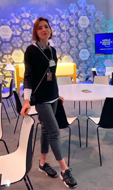 Внутренний павильон форума: человек с бейджем стоит у декоративной стены Всемирного экономического форума; вокруг столы, стулья и информационные панели мероприятия.