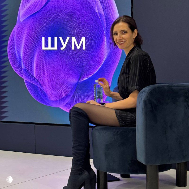 Автор канала сидит в кресле на сцене в черном платье и высоких сапогах, улыбается на фоне большого экрана с надписью «ШУМ».