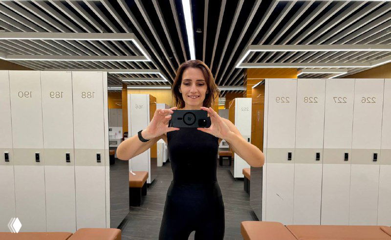 Девушка в спортивной одежде делает селфи в раздевалке фитнес‑клуба на фоне шкафчиков и зеркала с мягким светом.
