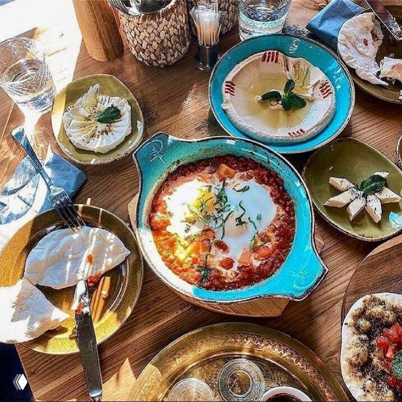 Вид стола с ливанскими мезе: тарелки с хумусом, баклажанными закусками, супом, лепёшками и десертом на деревянной поверхности в тёплом свете.