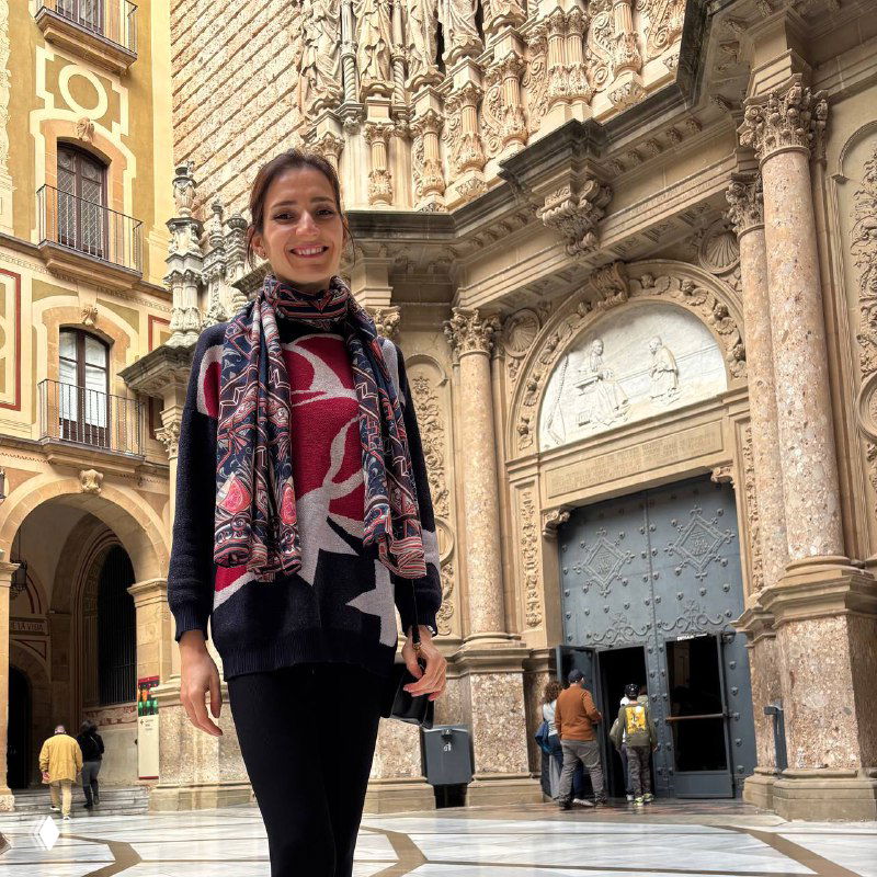 Автор у входа в собор Монсеррат: женщина в шарфе и свитере на фоне богато украшенного каменного портала и массивных дверей монастыря.