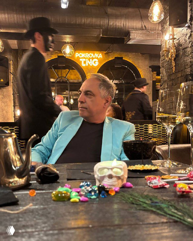 Мужчина в светлом пиджаке за столом в ресторане с тематическим декором и рассыпанными мелкими предметами — сцена ужина-перформанса.