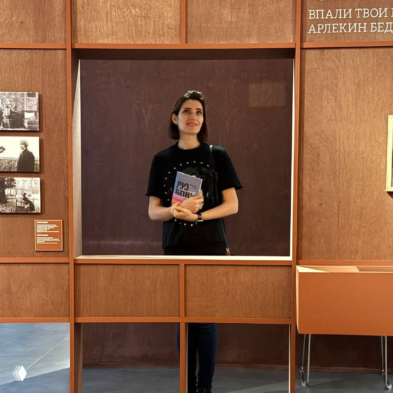 Женщина в зеркальной рамке инсталляции Центра «Зотов», держащая книгу; вокруг видны панно и фотоматериалы выставки «Любовь в авангарде».