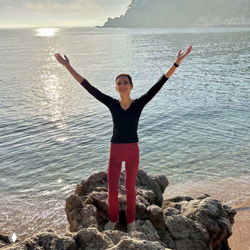 Женщина в чёрном топе и красных брюках стоит на скалистом берегу у моря с поднятыми руками; солнце отражается в воде, прогулка как часть здоровья.