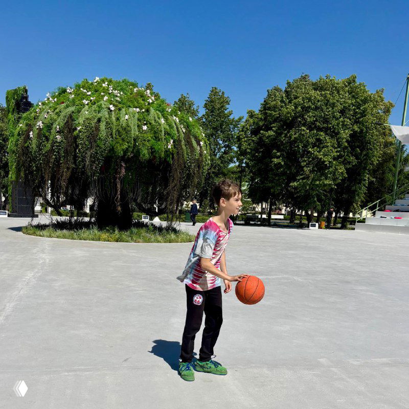 Ребёнок с баскетбольным мячом и рюкзаком на просторной городской площади у зелёных деревьев, тёплый ясный летний день, уличные активности.
