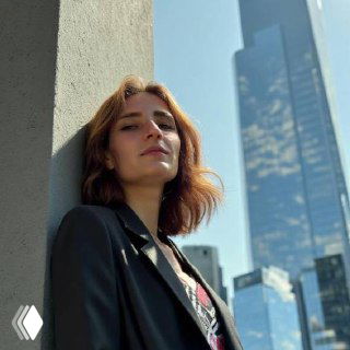 Портрет молодой женщины в пиджаке, прислонившейся к колонне на фоне стеклянных небоскрёбов и неба; крупный городской кадр, дневное освещение.