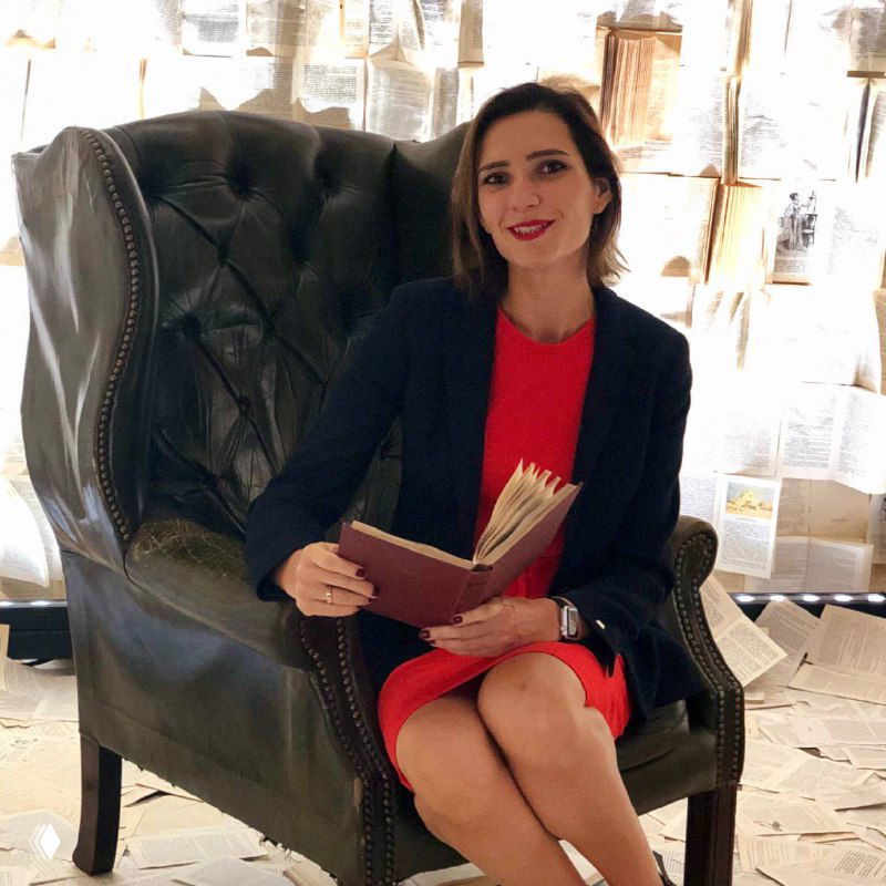 Женщина в красном платье сидит в кресле с раскрытой книгой на фоне стен, украшенных развешанными страницами — атмосферный литературный портрет.