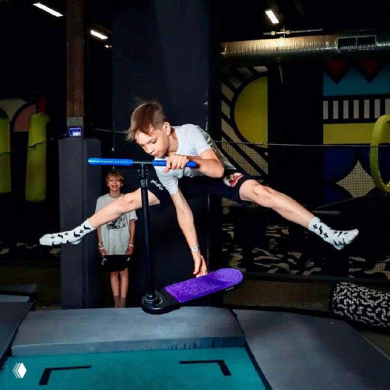 Мальчик в прыжке на батуте в детском развлекательном центре; видны элементы площадки, другие дети и мягкие маты вокруг.