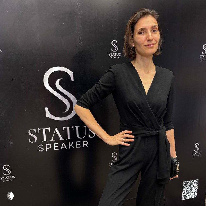 Женщина в чёрном костюме позирует у брендированной фотозоны «Status Speaker» на закрытом мероприятии; портрет в полный рост на фоне баннера с логотипом