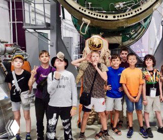 Группа детей-подростков во время экскурсии в инженерном музее на фоне авиационной техники; улыбающиеся мальчики и девочки в повседневной одежде, музейная обстановка.