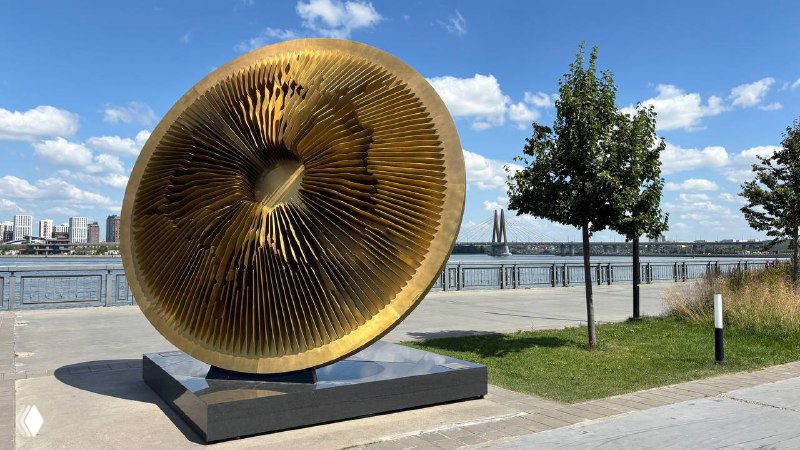 Большая металлическая и художественная круглая скульптура на прогулочной набережной Казанки; за ней река, деревья и городской горизонт.