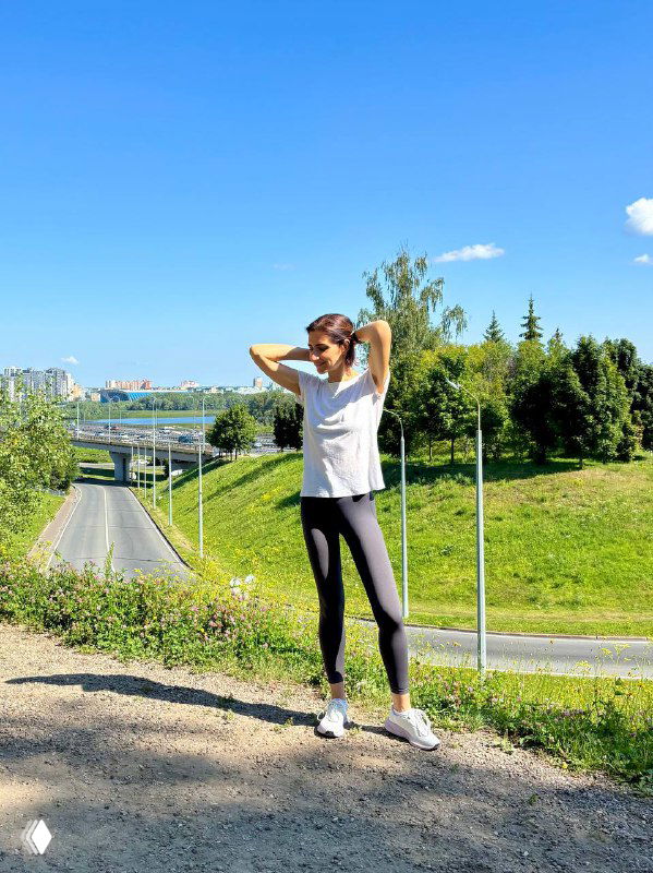 Автор позирует стоя на дорожке у пригорка в спортивной одежде; на заднем плане деревья, зелень и фрагменты городской застройки под голубым небом.