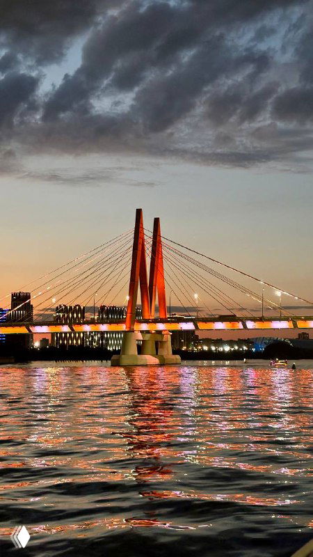 Вечерняя фотография вантового моста, подсвеченного огнями, с отражением в реке; небо в драматических облаках на закате.