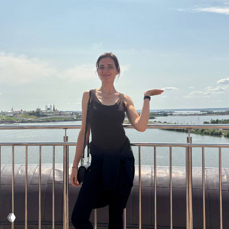 Автор позирует у перил на набережной Казани на фоне реки и панорамы города в ясный летний день, видны небоскрёбы и небо.