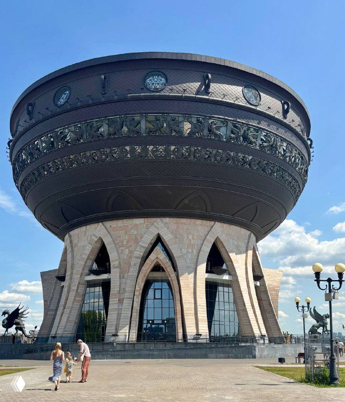 Здание Центра семьи «Казан» в форме огромной чаши с орнаментом по борту, расположенное у набережной и освещённое дневным солнцем.