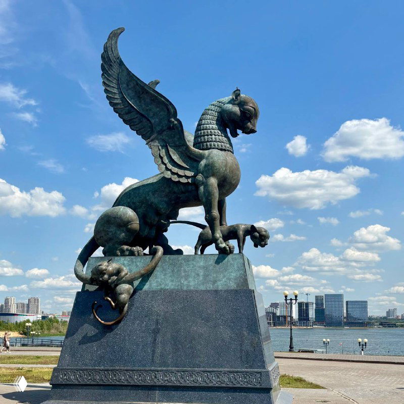 Крупный план бронзовой скульптуры зиланта — крылатого дракона на постаменте, установленного на набережной Казани на фоне неба.
